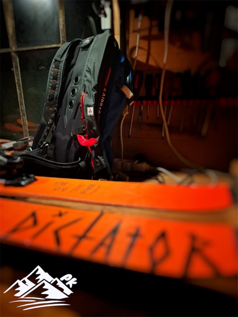 „Lavinový batoh airbag připravený na freeride, opřený v servisu vedle lyží připravených na ruční voskování. Výbava pro bezpečný pohyb v horách – servis a příprava lyží na Prostekram.cz.“